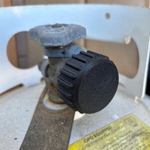 May include: A propane tank valve assembly featuring a gray metal handle and a large, circular black plastic knob with a textured, ridged edge. The assembly is mounted on a metallic tank with a yellow warning label that reads OPD EQUIPPED.
