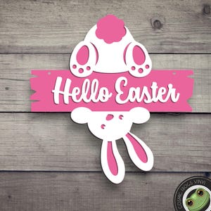 May include: A pink wooden sign with the text "Hello Easter" in white script. The sign features a white bunny design with pink accents, including the bunny's tail and ears. The sign is set against a wooden background.