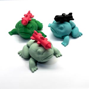 May include: Three frog figurines, each with a different colored missile on their head. The frogs are green, teal, and light green. The missiles are red, black, and pink.