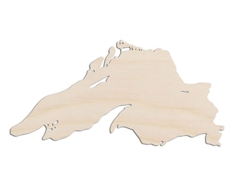 May include: A light-colored, unfinished wooden cutout shaped like the geographic outline of Lake Superior. The piece features a smooth, flat surface with visible natural wood grain patterns and irregular, jagged edges that define the shoreline.