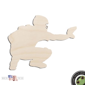 May include: A light-colored, unfinished wooden cutout in the shape of a baseball catcher in a crouched position, wearing a helmet and chest protector, with one arm extended forward as if catching a ball, featuring a smooth, natural wood grain.