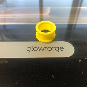 May include: A bright yellow plastic ring, likely a plug or spacer, with a smooth surface and a circular hole in the center. The ring is sitting on a gray surface with the word "glowforge" printed on it.