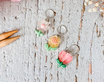 May include: Three flower-shaped stitch markers with silver rings. The markers are pink, yellow, and peach with green leaves.