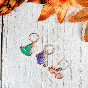 May include: Three Halloween-themed charms with gold-colored rings. The charms include a green witch hat with gold stars, a purple ghost, and a pink bat. The charms are on a white, distressed wooden surface with fall foliage.