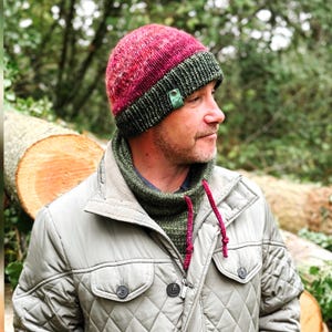 May include: A handmade knit beanie in shades of burgundy and green, with a matching green neck warmer. The beanie has a tag attached. The person is wearing a light gray quilted jacket.