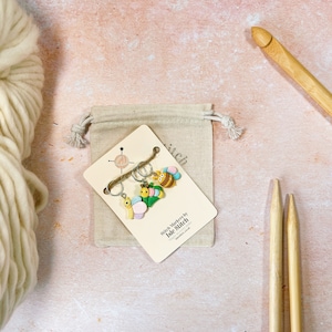 May include: A collection of stitch markers with a butterfly, snail, and bee design, presented on a card with the text "Stitch Markers by Isle Stitch". The markers are on a small beige drawstring bag, with a large wooden crochet hook and knitting needles.