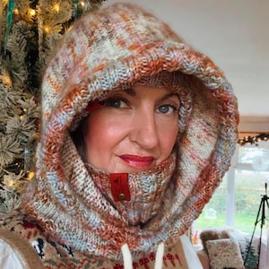 May include: A hooded cowl in a mix of orange, cream, and blue yarns. The knitted cowl has a drawstring and a small red leather tag with a button closure. The hood is up, and the cowl is around the neck.
