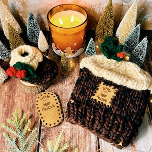 May include: Two knitted Christmas pudding ornaments with holly and berry embellishments. The ornaments are brown and white with a cream colored top. The ornaments have a brown leather tag with the text "Little Pudding" on them.