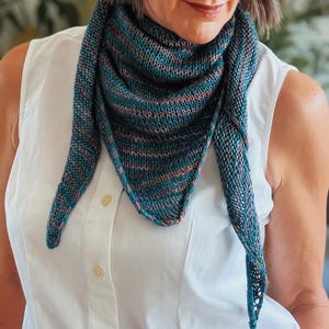 May include: A triangular knit scarf featuring a variegated color palette of deep teal, dark blue, and soft peach tones. The scarf is constructed with a textured stockinette stitch pattern and is draped around the neck over a white button-down shirt.