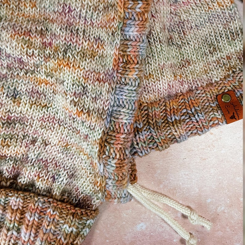 May include: Close-up of a hand-knitted hat in a speckled pattern of beige, orange, pink, and purple. The hat has a drawstring and a small leather tag with a logo. The texture of the knitwear is visible.