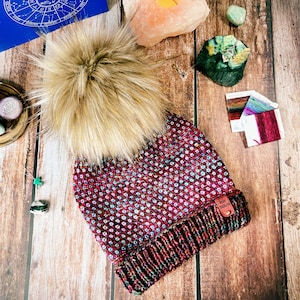 Könnte beinhalten: Eine rot-weiß gefleckte Strickmütze mit einem Kunstfellbommel oben. Die Mütze hat ein braunes Leder-Etikett mit dem Text "The Stitch" darauf.