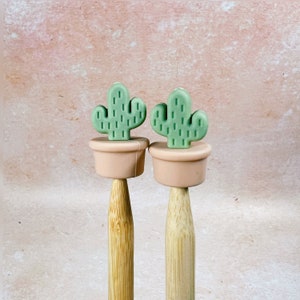 May include: Two wooden knitting needles with green cactus-shaped silicone toppers. The cactus toppers are in pink pots.