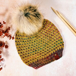 Op de afbeelding: Een gele, roze en groene gebreide beanie met een bruine nep bont pompon. De beanie heeft een bruin leren label met het woord "Handmade" erop.