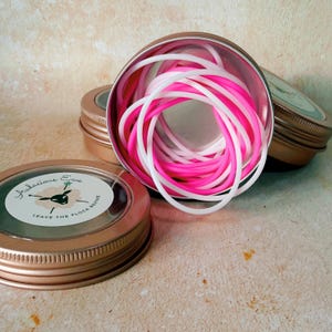 May include: A collection of pink and white silicone bracelets inside a small, open, metallic tin. The tin is surrounded by other similar tins with lids. The lids have a label with the text "Audacious Ewe" and "Leave the Flock Behind".
