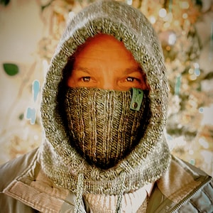 May include: A knitted hood and face covering in shades of gray and green. The hood is pulled up, with the face covering extending to the neck. A small green tag is attached to the face covering. The background is blurred.