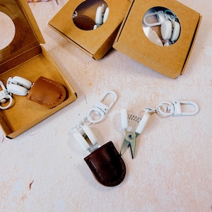May include: Two brown leather-effect scissor sheaths with white metal clips and white swivel hooks. One sheath is inside a square brown cardboard box with a circular cutout, and two others are displayed outside with one open to reveal metal scissors.