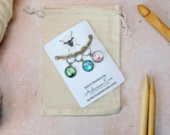 May include: Set of three stitch markers with floral designs. The markers are silver with clear glass tops. The designs are a pink floral pattern, a blue and green floral pattern, and a green floral pattern. The markers are on a white card with the text "Stitch Markers by Audacious Eve Leave the Flock Behind audaciousweve.etsy.com".