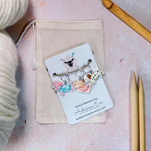 May include: A set of three dog-shaped stitch markers on a white card with the text "Stitch Markers by Audacious Ewe". The markers are attached with silver rings and are in pink, white, and brown colors. A beige drawstring bag and knitting needles are also in the image.