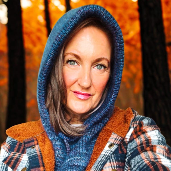 May include: A woman wearing a blue knit hood and a plaid flannel jacket with a brown sherpa collar. The jacket has a button closure. The background is a blurred view of trees with orange leaves.