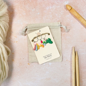 May include: A set of three colourful stitch markers with a rainbow, flower, and cactus design. The markers are made by Isle Stitch and are packaged in a small white drawstring bag.