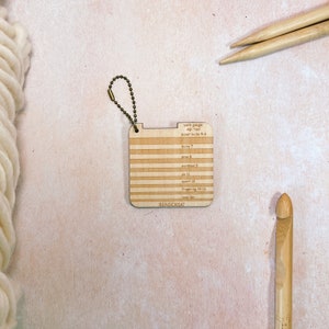 May include: A wooden yarn gauge with a metal keychain. The gauge has a ruler with markings for different yarn weights, including super bulky, bulky, worsted, dk, sport, fingering, and lace.