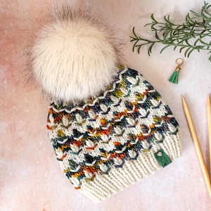 Könnte beinhalten: Eine Strickmütze mit einem großen cremefarbenen Kunstfell-Bommel und einem mehrfarbigen Muster aus orangefarbenen, grünen und dunkelblauen Stichen auf cremefarbener Basis. Die Mütze hat ein geripptes cremefarbenes Bündchen mit einem kleinen grünen Etikett.