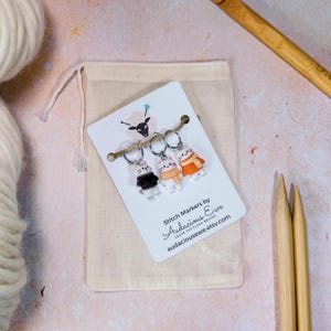 May include: A set of three cat-shaped stitch markers on a white card with the text "Stitch Markers by Audacious Ewe". The markers are attached to a small beige drawstring bag, with knitting needles and yarn in the background.