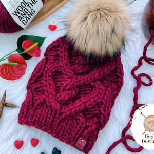 Könnte beinhalten: Eine burgunderfarbene Strickmütze mit einem großen Zopfmuster und einem Kunstfellbommel oben. Die Mütze liegt auf einer weißen, flauschigen Oberfläche mit einer Wollknäuel, Stricknadeln und kleinen herzförmigen Dekorationen.
