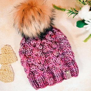 Könnte beinhalten: Eine rosa und lila gestrickte Mütze mit einem großen, flauschigen Bommel. Die Mütze hat ein Zopfmuster und ein Leder-Etikett mit dem Text "Love, Knit & Co."