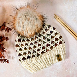 May include: Cream-colored knit hat with a diamond pattern and a large, fluffy, multi-toned faux fur pom-pom. The hat has a small leather tag and is next to wooden knitting needles. A cozy winter accessory.