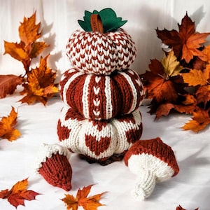 May include: A stack of three knitted pumpkins in red and white patterns, with a smaller knitted acorn on either side. The pumpkins are arranged from largest to smallest, with the top pumpkin featuring a brown tag that reads "MOURITE".
