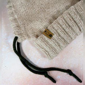 May include: A beige knitted hat with a ribbed cuff and dark green drawstrings. A small brown leather tag with text and a button is sewn onto the cuff. The hat is made of a soft, textured yarn.