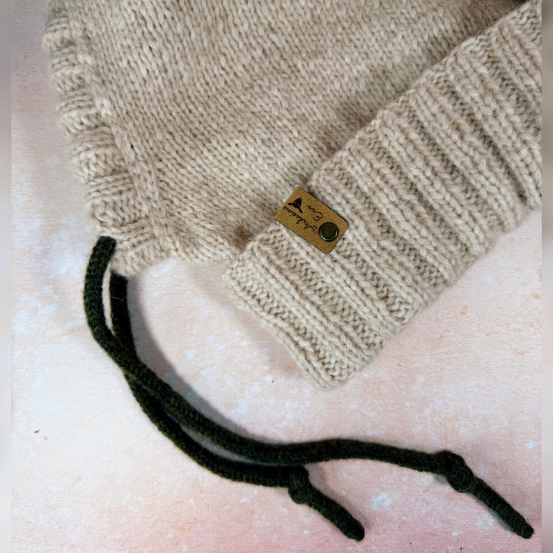 May include: A beige knitted hat with a ribbed cuff and dark green drawstrings. A small brown leather tag with text and a button is sewn onto the cuff. The hat is made of a soft, textured yarn.
