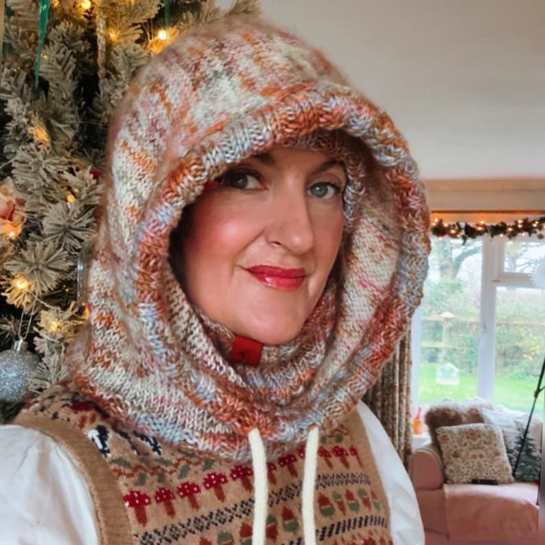 May include: A hooded cowl in a mix of orange, brown, and blue yarns. The cowl has a drawstring and is worn over a patterned sweater vest. The knitted hood is a cozy accessory for cold weather.