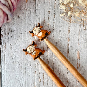 May include: Two wooden knitting needles topped with brown and cream Highland cow charms. The charms have black horns and eyes. The needles are set against a weathered, white-painted wooden surface, with a ball of pink yarn in the background.