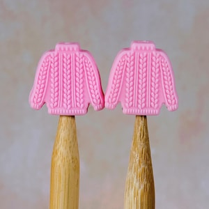 May include: Two pink sweater-shaped erasers with cable knit detailing are mounted on wooden pencils. The erasers are a vibrant pink color and have a textured surface. The pencils are made of light-colored wood.