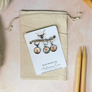 May include: A set of three stitch markers with fox illustrations, attached to a white card with the text "Stitch Markers by Audacious Ewe". The markers are in a beige drawstring bag, with two wooden knitting needles.