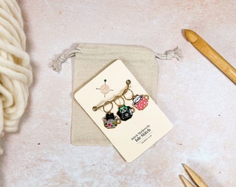 May include: A set of three stitch markers shaped like teacups, each with a unique design, attached to a cream-colored card. The card is inside a small beige drawstring bag. The text "Stitch Markers by Isle Stitch" is visible on the card.