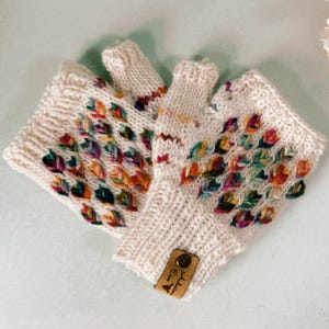 May include: A pair of white fingerless gloves with a colorful embroidered design. The gloves have a brown leather tag with the text "Soulful Stitches" on it.