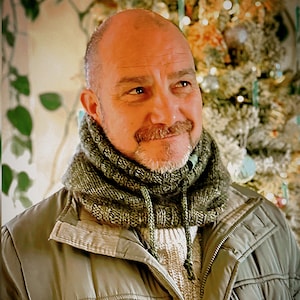 May include: A man wearing a green knitted cowl neck warmer with a drawstring. He is also wearing a light-colored sweater and a green jacket. The background is blurred, with a Christmas tree visible.