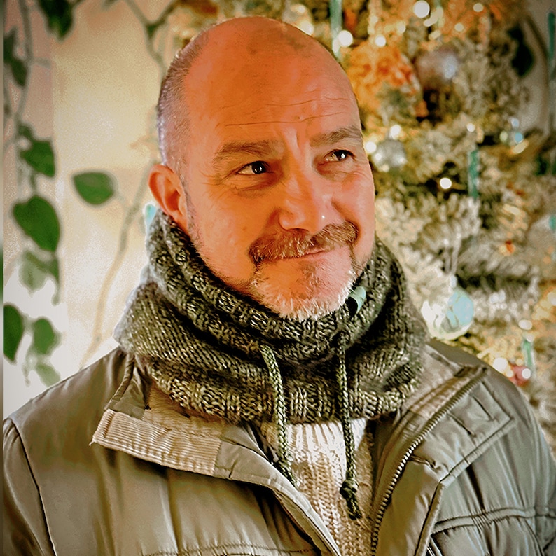 May include: A man wearing a green knitted cowl neck warmer with a drawstring. He is also wearing a light-colored sweater and a green jacket. The background is blurred, with a Christmas tree visible.