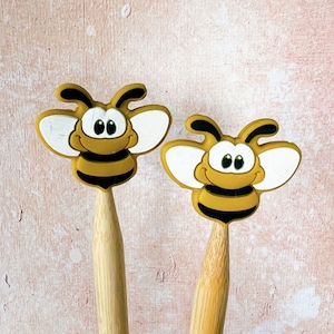 May include: Two wooden knitting needles with cute cartoon bee toppers. The bees are yellow and black with white wings and smiling faces.