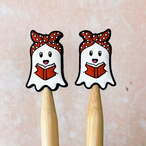 May include: Two white ghost-shaped pencil toppers with black outlines, each wearing a red polka-dot headband and holding a red book. The pencils are made of light-colored wood, and the background is a soft pink.