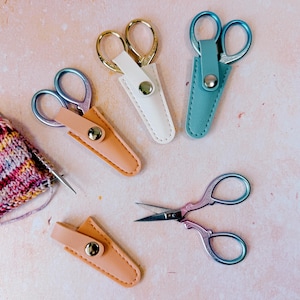 May include: Three pairs of small metal scissors with iridescent blue and purple handles, each stored in a protective faux leather sheath in peach, white, or teal. One pair of scissors is shown outside its peach sheath, alongside a piece of knitting.