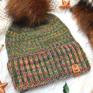 Könnte beinhalten: Eine Strickmütze mit einem melierten Garnmuster in Grün, Pink und Gelb, einem gerippten Umschlag und einem großen braunen Kunstfell-Bommel. Am Umschlag befindet sich ein kleines rechteckiges Etikett aus beigem Leder mit dem Aufdruck Isle Stitch.