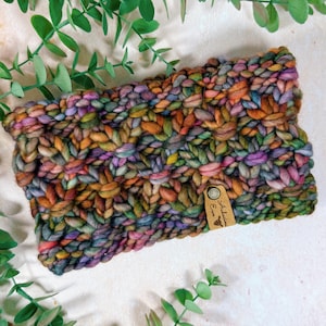 Pode incluir: Uma faixa de cabeça de malha grossa multicolorida, com tons de laranja, verde, roxo e castanho. A faixa de cabeça tem uma etiqueta que diz "Artisan Eve". O artigo é feito de fio grosso e texturizado.