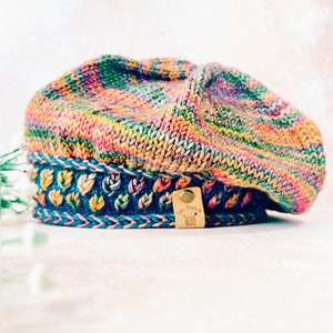 Könnte beinhalten: Eine bunte gestrickte Baskenmütze mit blauem und rosafarbenem Besatz und einem kleinen Lederanhänger mit der Aufschrift "Julie Stitch".