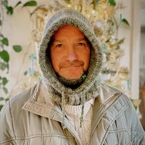 May include: A knitted hood in shades of green and gray, with a drawstring closure. The hood is worn over a light-colored sweater and a green jacket. The background features a blurred, festive arrangement with lights and greenery, suggesting a winter theme.