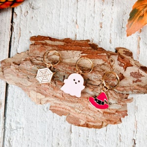 May include: Three Halloween-themed charms with gold-colored rings. The charms include a white spiderweb, a pink ghost, and a red witch hat with a black band. The charms are displayed on a piece of tree bark.