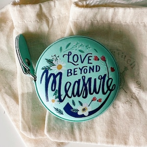 May include: A light blue round measuring tape with white flowers and red hearts. The tape reads "Love Beyond Measure".